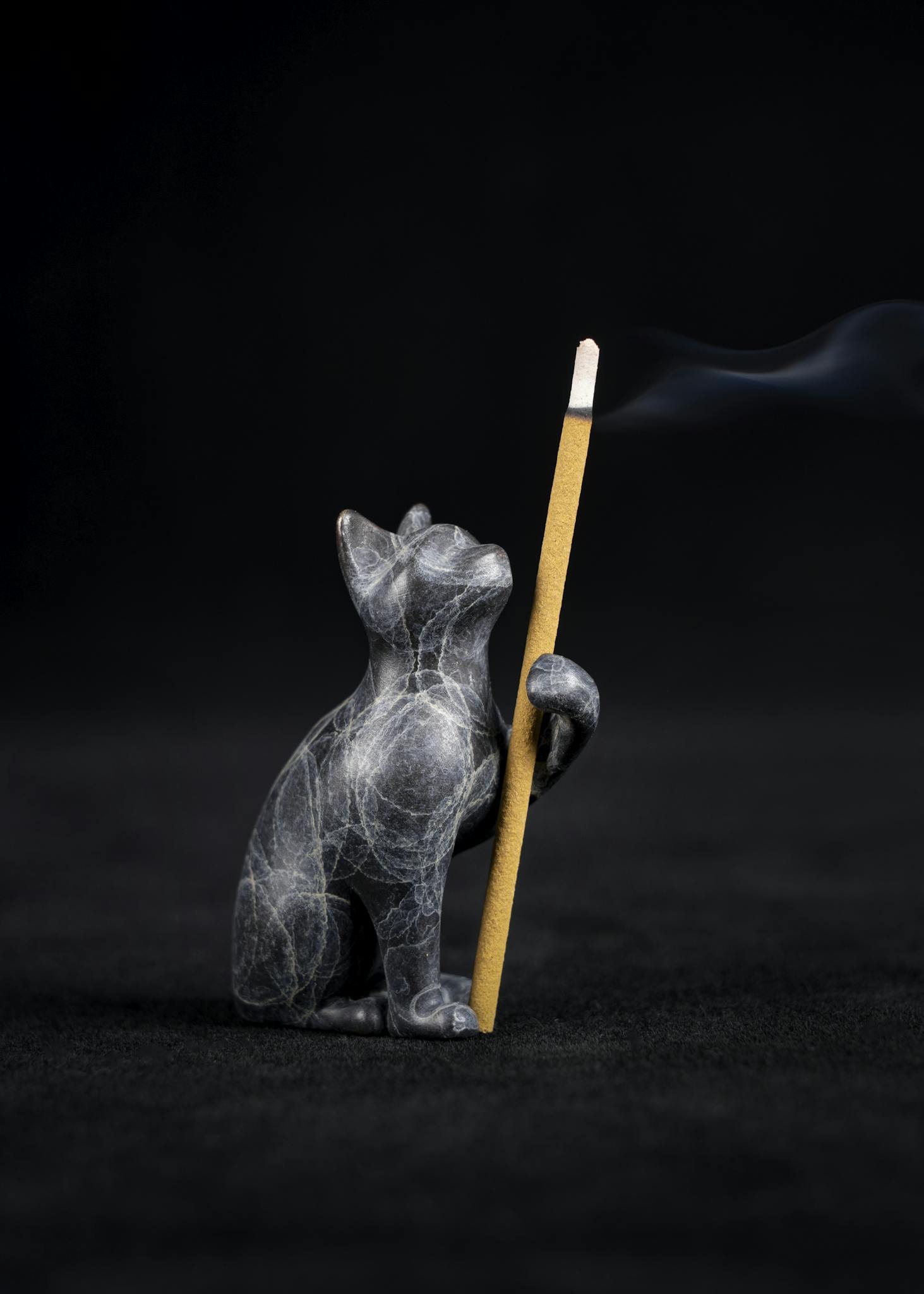 Marble cat figurine gracefully holding a burning incense stick, set against a dark background with visible smoke.